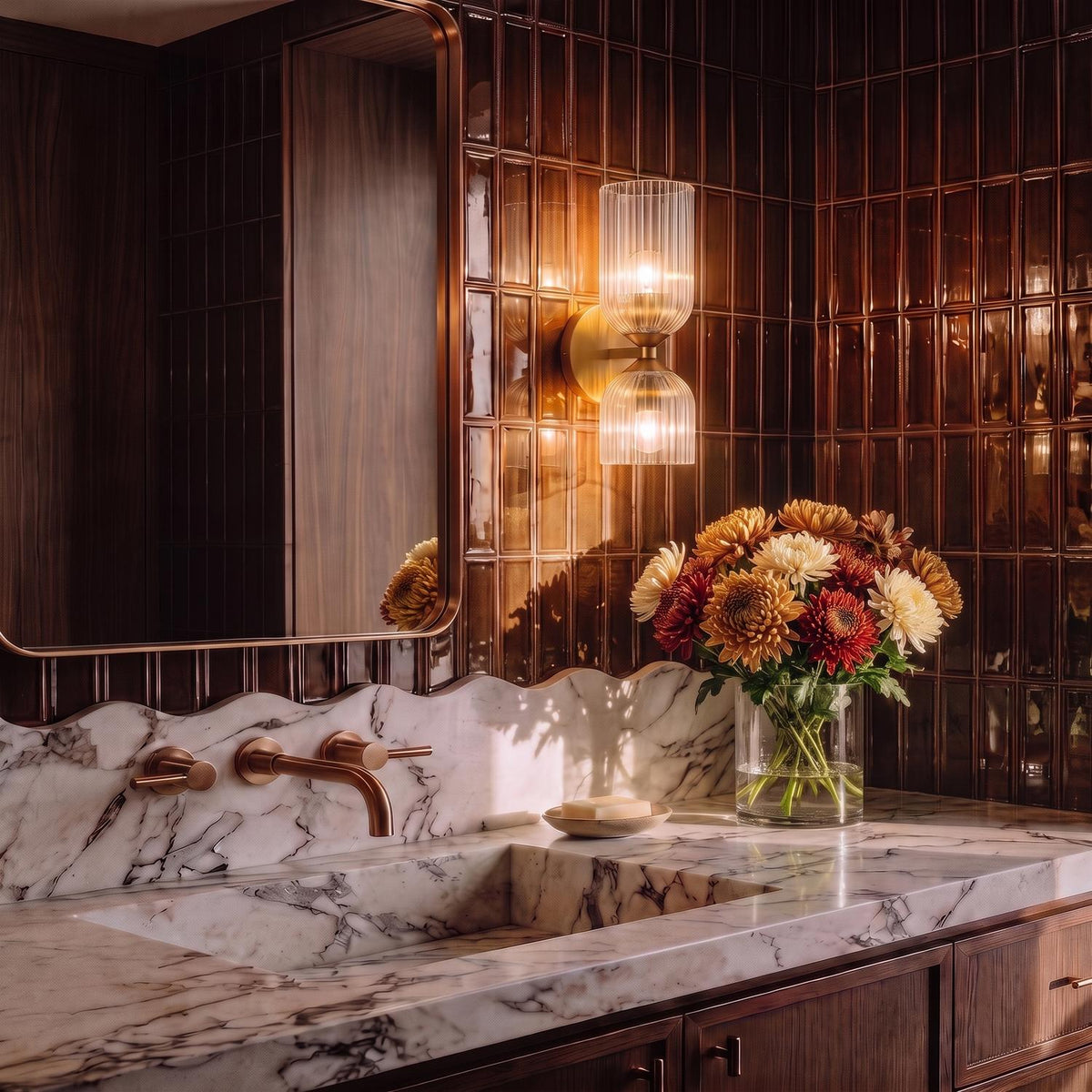A mid-century bathroom featuring an Elle Wall Sconce, Brushed Brass and Clear Glass mounted on brown vertical tiles above a marble vanity and integrated sink.
