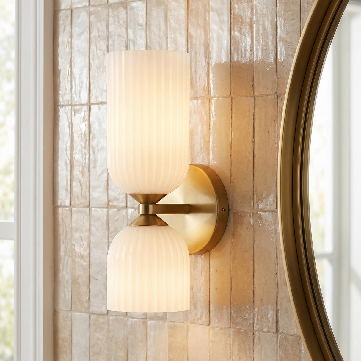 A transitional bathroom with an Elle Wall Sconce, Brushed Brass and Milk Glass mounted on a textured beige tile wall beside a round gold mirror.