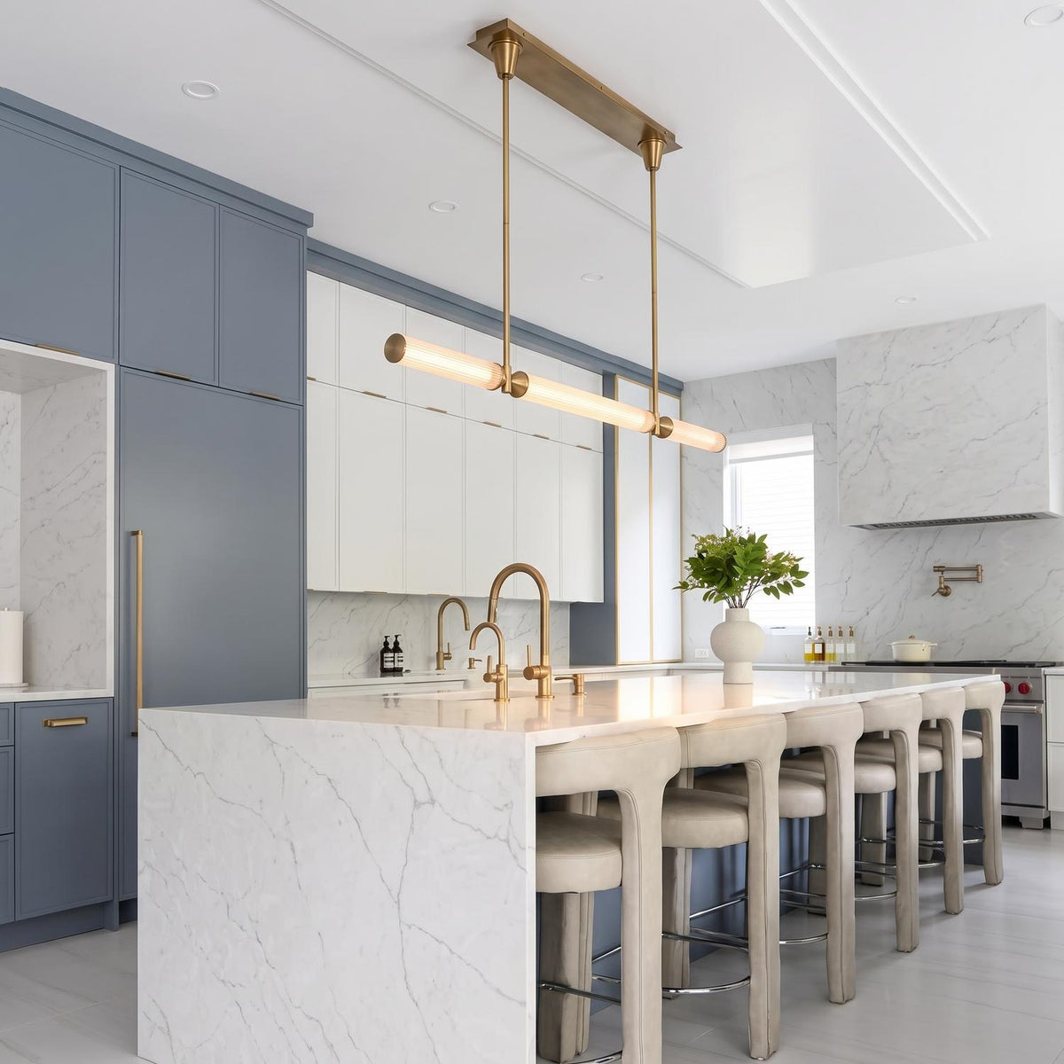 A modern kitchen featuring the Blain LED Linear Pendant 48", Vintage Brass with ribbed glass suspended over a marble waterfall island lined with beige barstools and blue cabinetry.