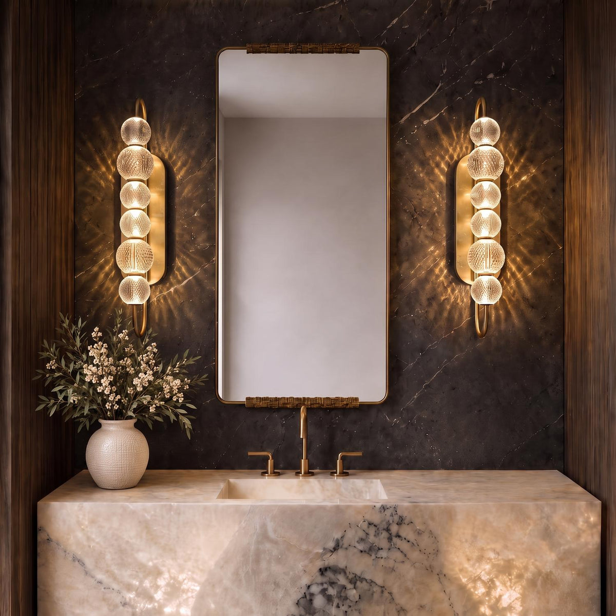 A modern bathroom features natural brass wall sconces with textured glass globes flanking a brass-framed mirror over an illuminated marble vanity counter and dark stone wall.