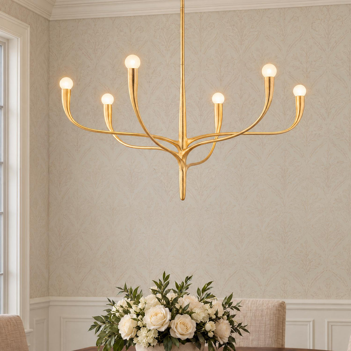 A transitional dining room featuring a gold leaf six-light chandelier suspended above a dining table decorated with a white floral centerpiece and subtle patterned wallpaper.