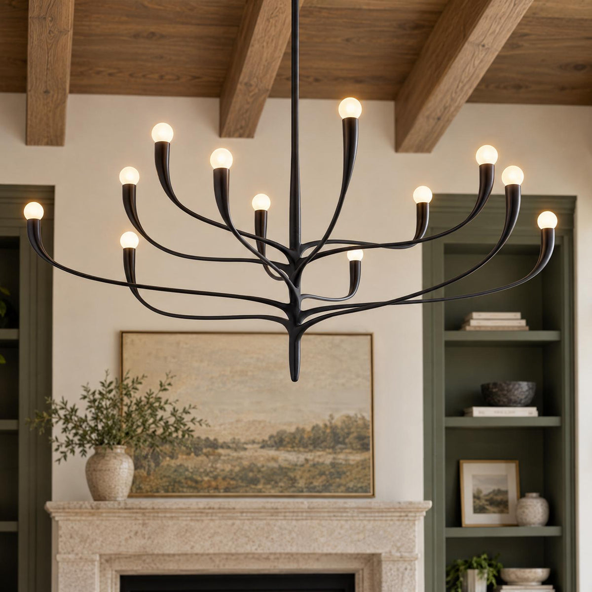 A transitional living room features an organic antique iron twelve-light chandelier suspended beneath a wooden beamed ceiling above a stone fireplace with green built-ins.