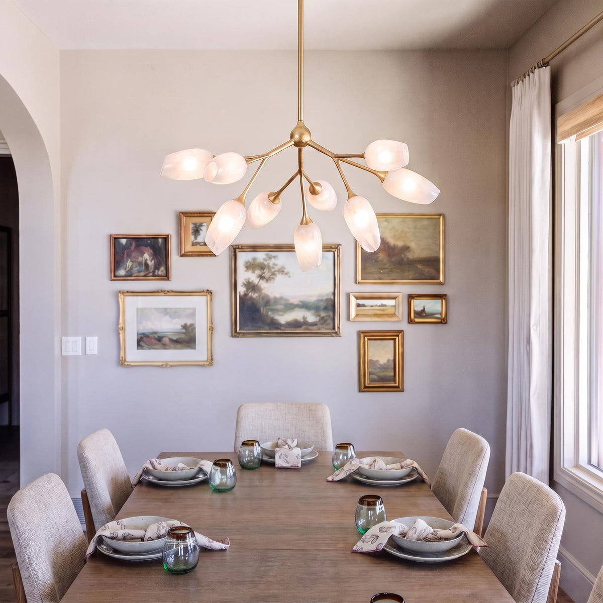 A transitional dining room features the Amara 9 Light Chandelier in gold leaf with art glass suspended over a wooden table before a framed gallery wall.