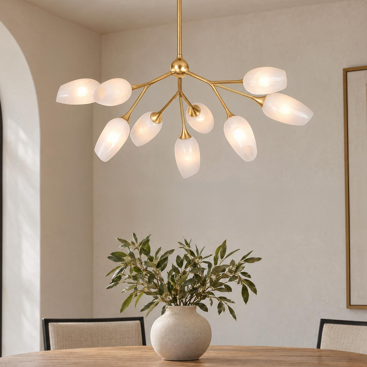 A contemporary dining room features the Amara 9 Light Chandelier 34", Gold Leaf with Art Glass suspended over a wood table topped with a ceramic vase.