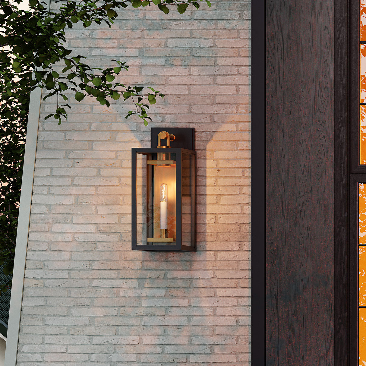 Orin outdoor medium wall light in bronze with brass accents is shown glowing during sunset against a white washed brick wall.