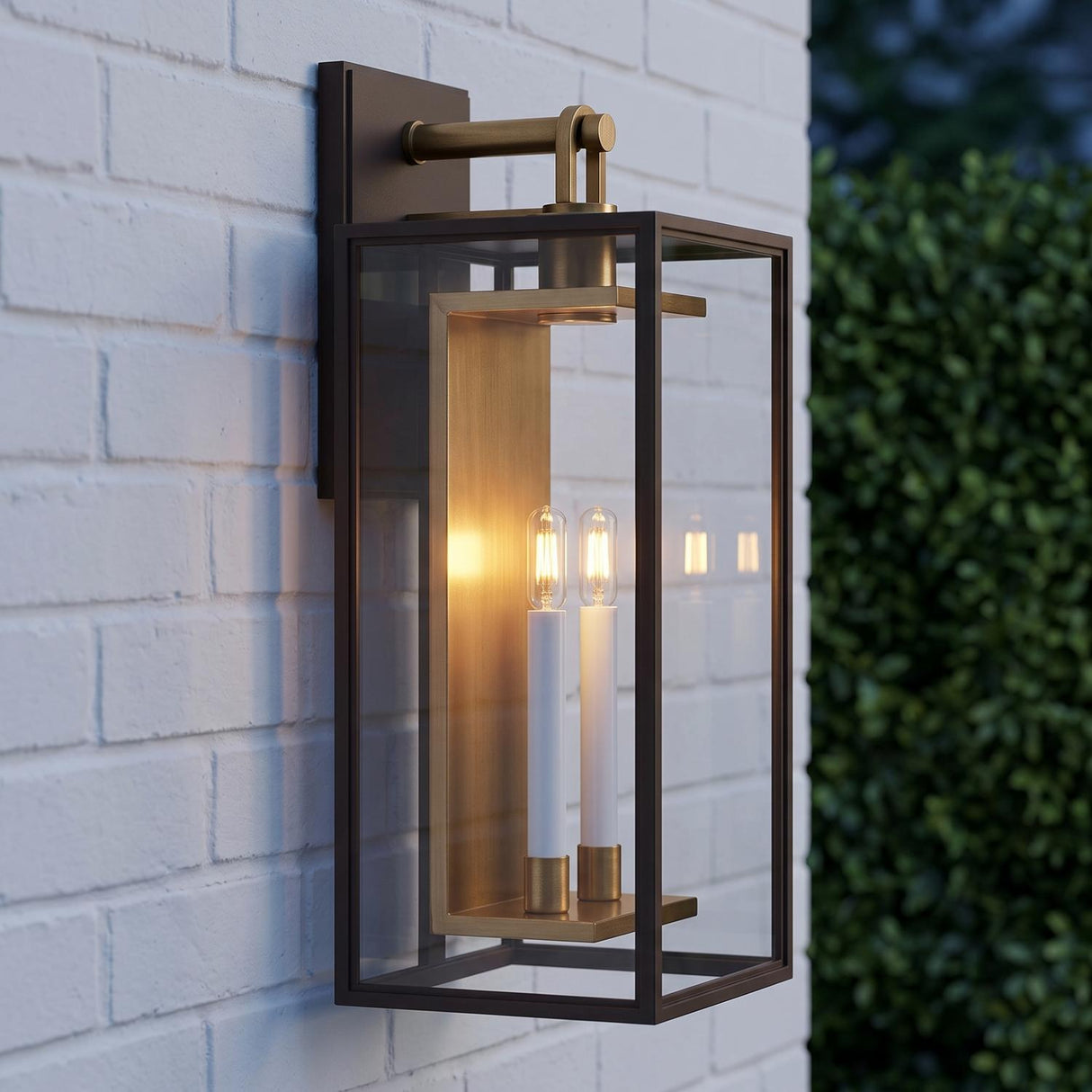 A modern farmhouse exterior featuring the Orin Outdoor Wall Light in bronze with vintage brass accents glowing on white brick beside green boxwood at twilight.