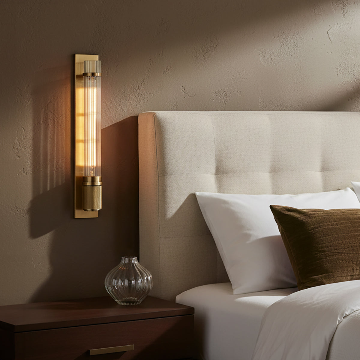 Bedroom scene with the Zeke Wall Sconce in Aged Brass finish, nightstand, and bed with pillows.