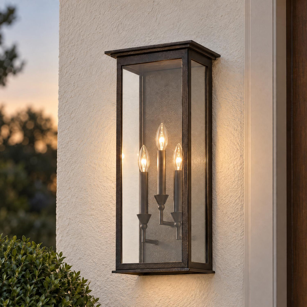 A transitional home entryway features an oiled bronze wall lantern glowing at dusk on a white stucco wall beside a dark wood door near green boxwood.