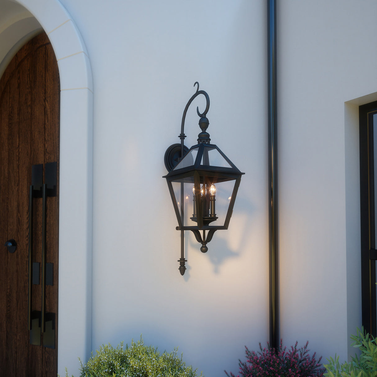 Decorative outdoor wall light fixture on a white wall with a wooden door and plants in the foreground.