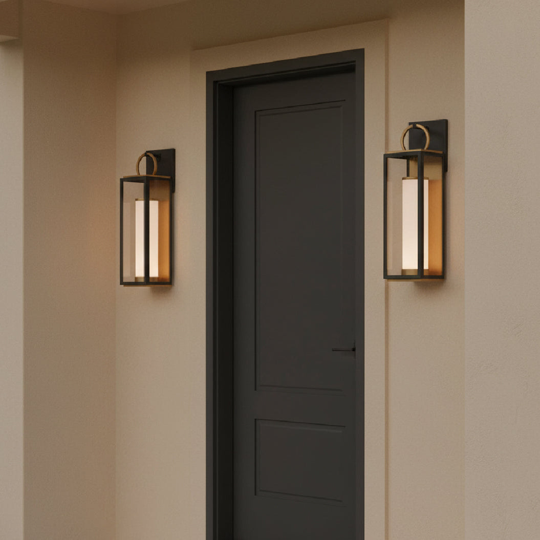Beige stucco walls with a dark door between two wall-mounted Waverly LED Outdoor Wall Lights, Medium 23" Black with Vintage Brass Accents.