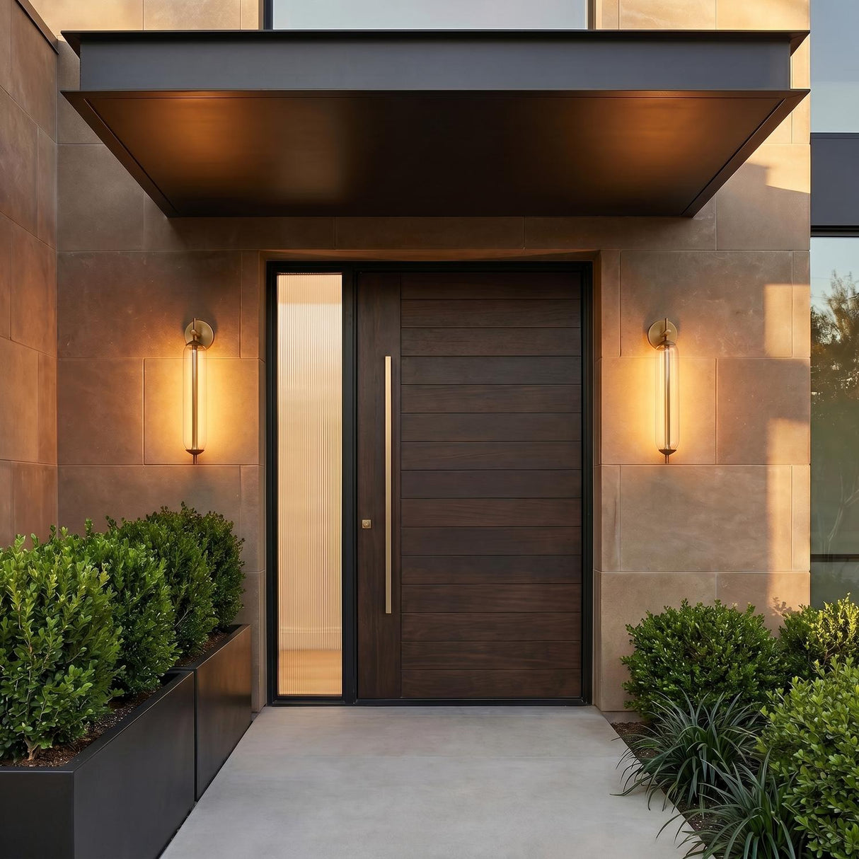A modern home entry with Marcy LED Outdoor Wall Light, 21" Vintage Brass sconces flanking a dark wood door on stone panels at twilight with boxwood hedges.