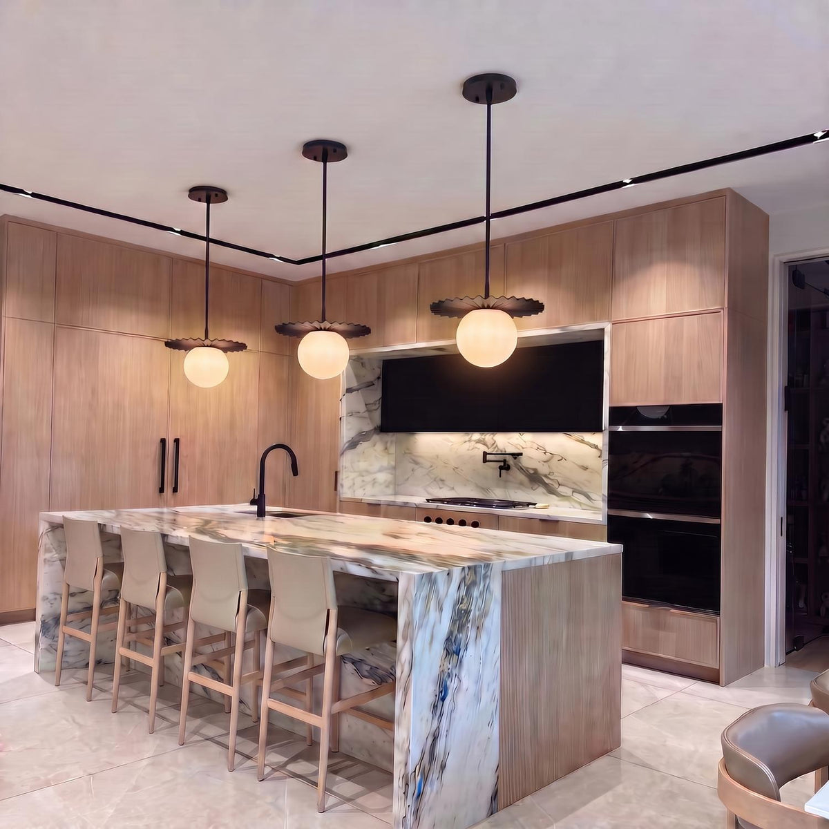A modern kitchen features three matte black globe pendants with opal glass shades hanging over a dramatic marble waterfall island surrounded by light wood cabinetry.