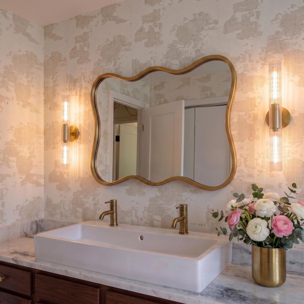 A transitional bathroom featuring two aged brass wall sconces with ribbed glass flanking a wavy gold mirror above a white vessel sink and marble countertop.