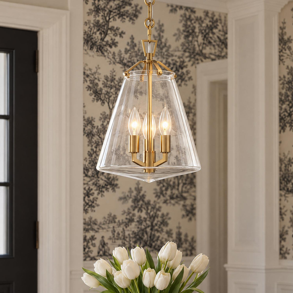 A transitional foyer features a brushed brass pendant light with a clear glass lantern shade suspended near black and white tree wallpaper and white tulips.