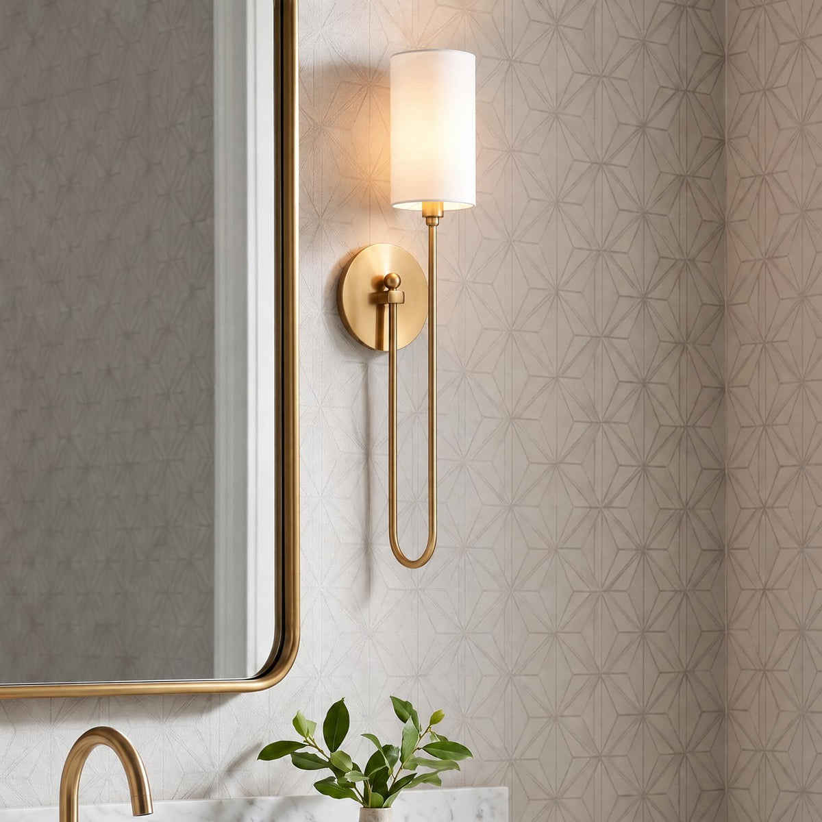 A transitional bathroom featuring an aged brass wall sconce with a white fabric shade mounted on geometric wallpaper beside a brass mirror above a marble countertop.