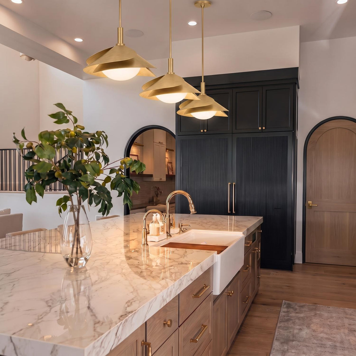 A transitional kitchen with three Lola Pendants in gold leaf with milk glass over a white marble island, featuring fluted black cabinetry and arched wood doors.
