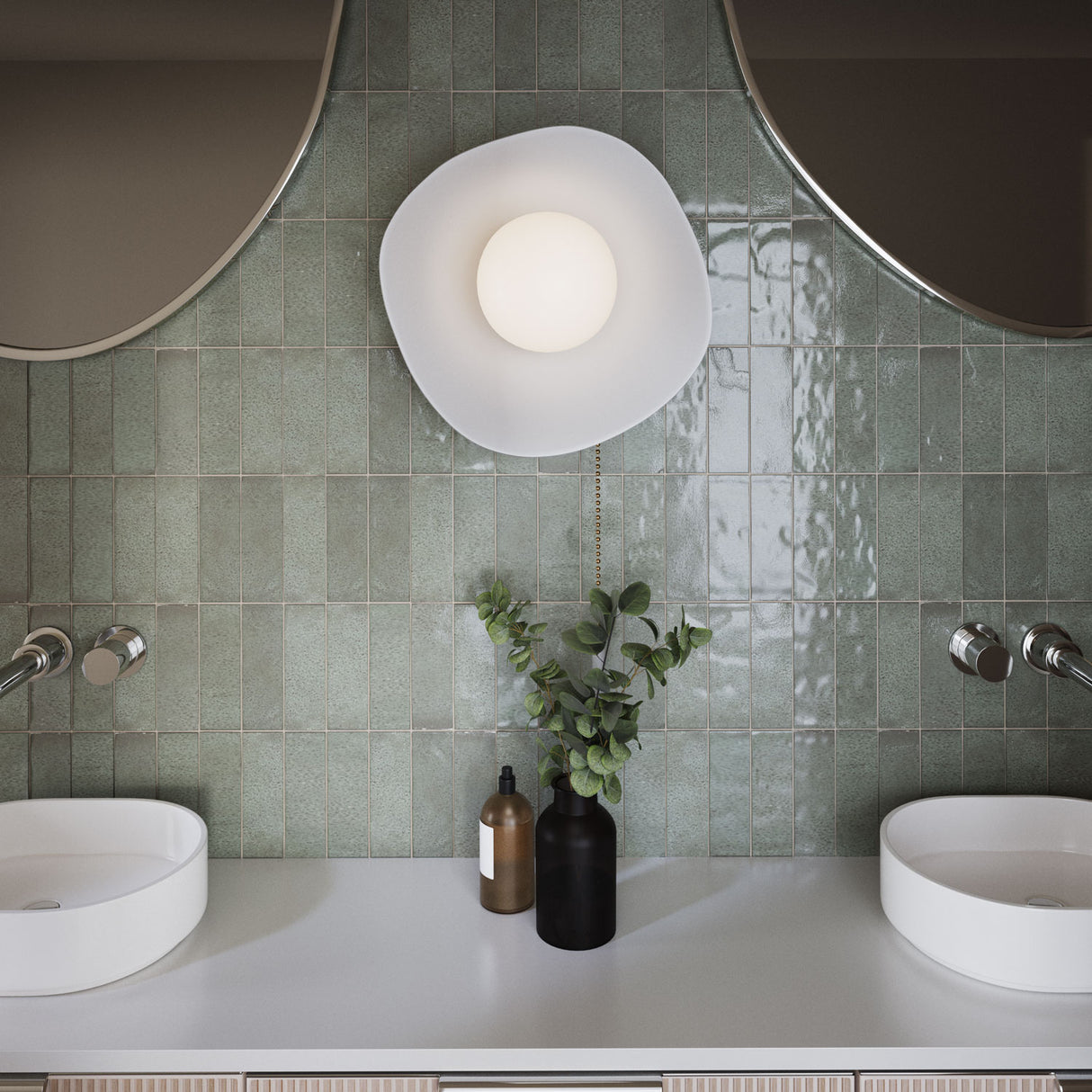 The Ulla Wall Sconce, White Ceramic with Brass Accents, is shown close-up in a modern bathroom with sage green tiles and chrome hardware.