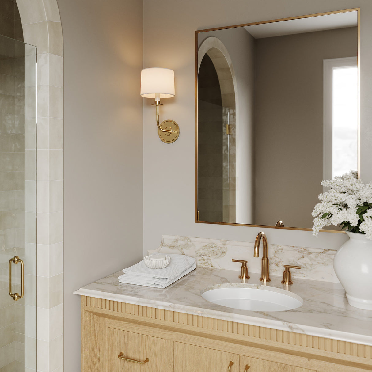 Marlowe Wall Sconce 14", Aged Brass finish with white fabric shade, is shown next to a brass vanity mirror in a bathroom.
