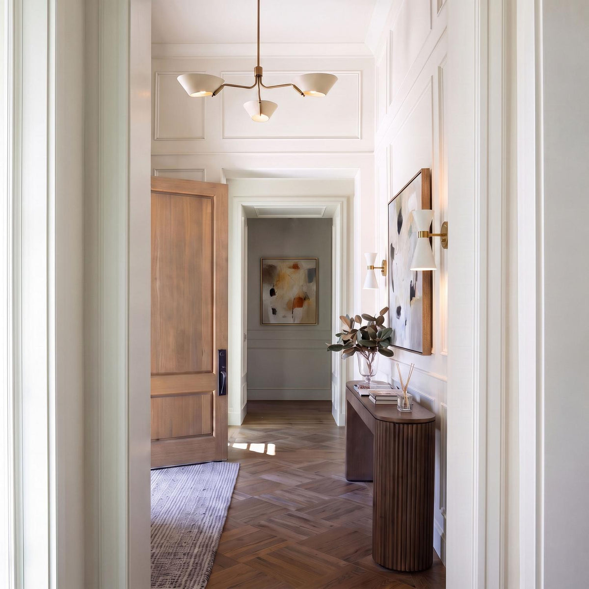 A transitional foyer with the York 3 Light Chandelier in patina brass and soft sand above herringbone wood floors, white paneled walls, and a fluted wood console.