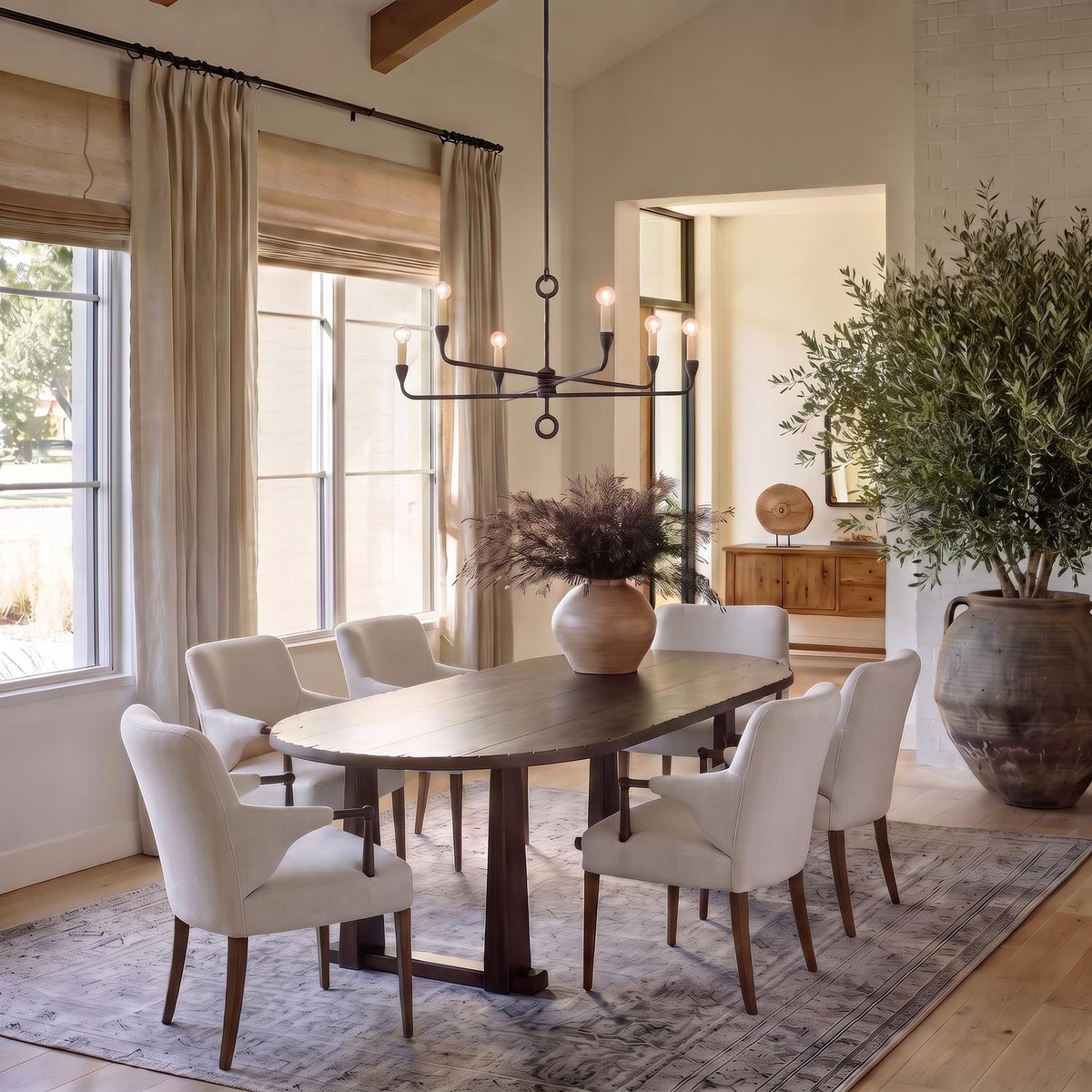 A transitional dining room featuring an antique iron six-light chandelier suspended over an oval dark wood dining table surrounded by cream armchairs under exposed wooden ceiling beams.