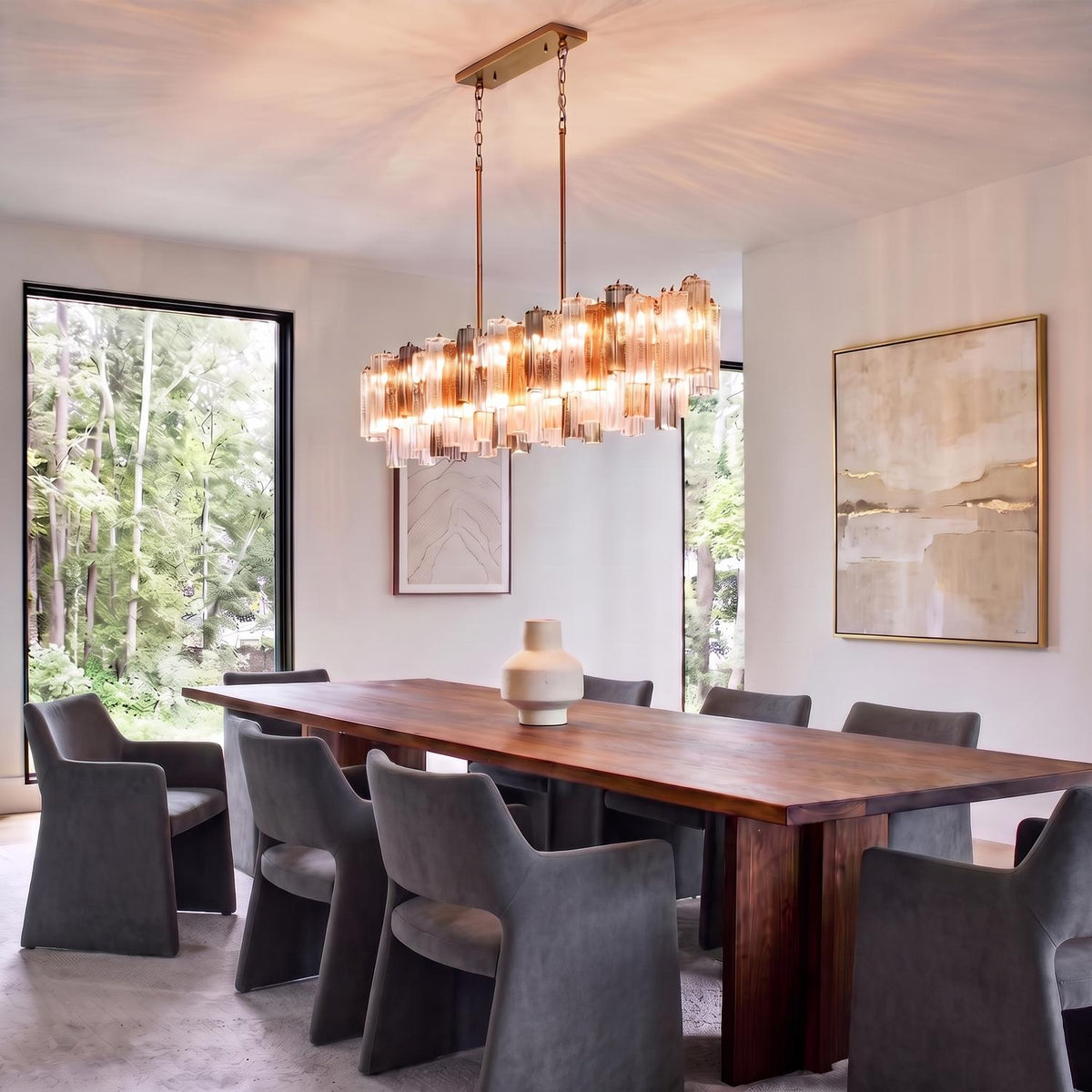 A contemporary dining room featuring a brass linear chandelier with gilded fog glass suspended over a long wood table and gray upholstered chairs.