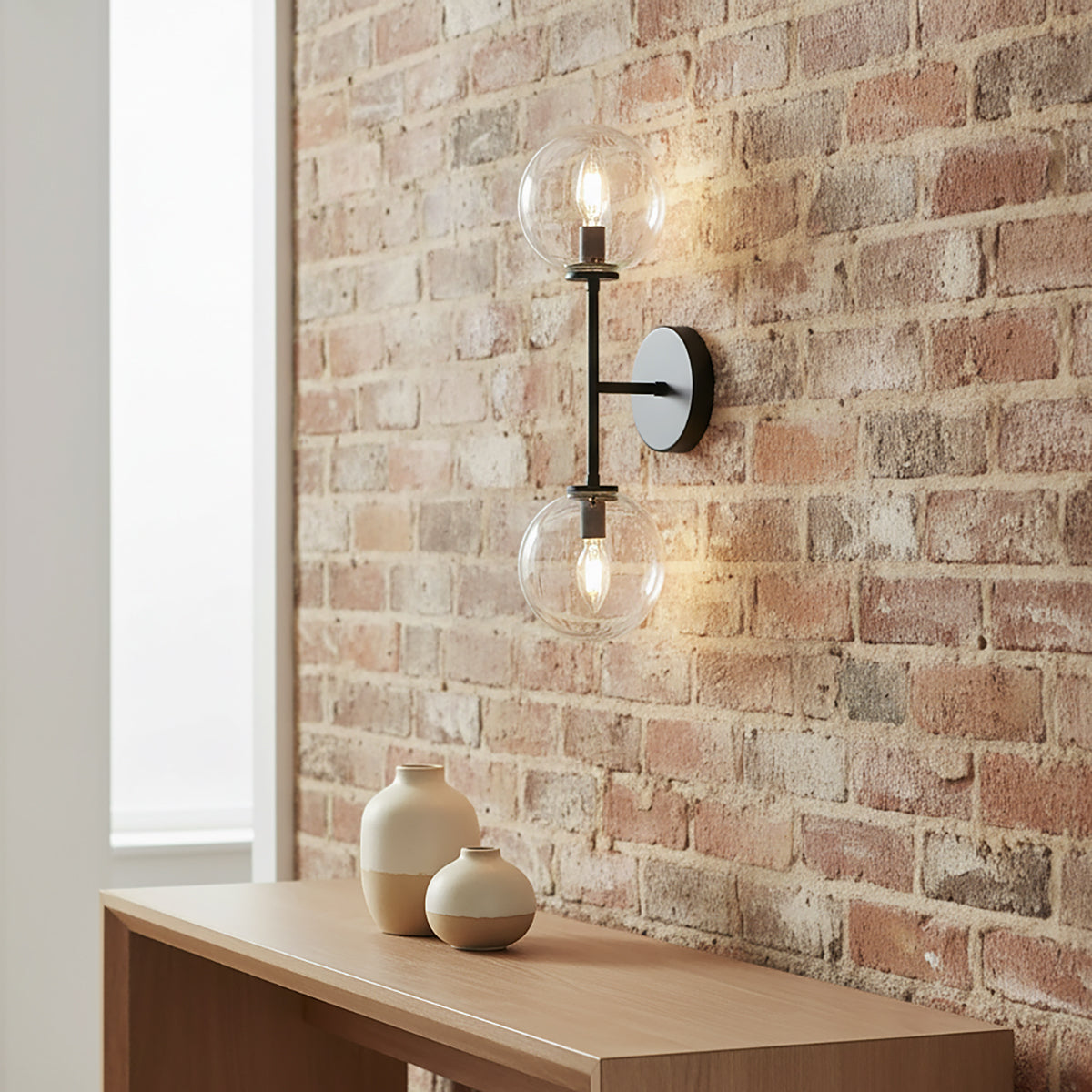 Gurt 2 Light Wall Sconce 20", Matte Black with clear glass on an exposed brick wall with a wooden shelf and decorative vases.
