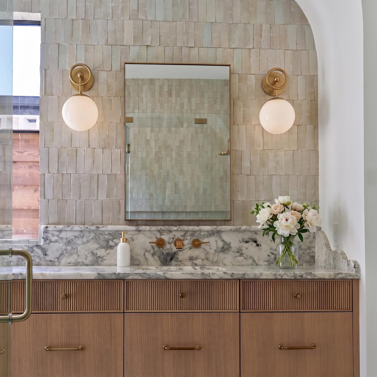 A transitional bathroom featuring two Tristen Wall Sconce 12 inch fixtures in brushed brass and opal glass flanking a mirror over a marble countertop and fluted cabinets.