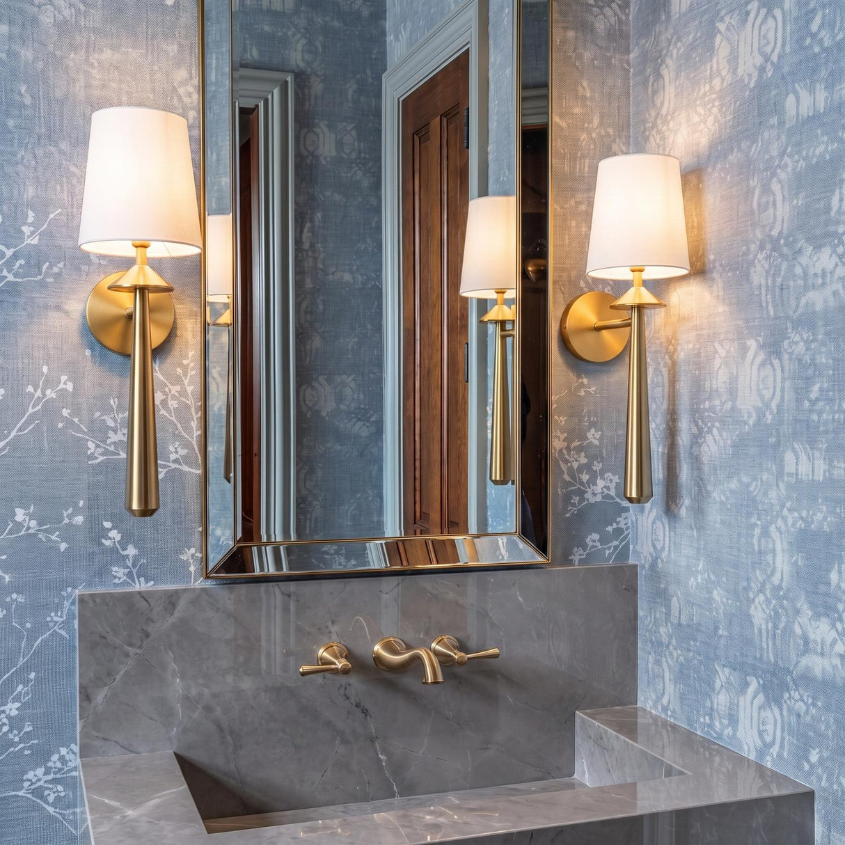 A transitional bathroom with two Sabina Wall Sconce 18-inch brushed brass fixtures featuring white linen shades flanking a gold framed mirror above a gray marble sink.
