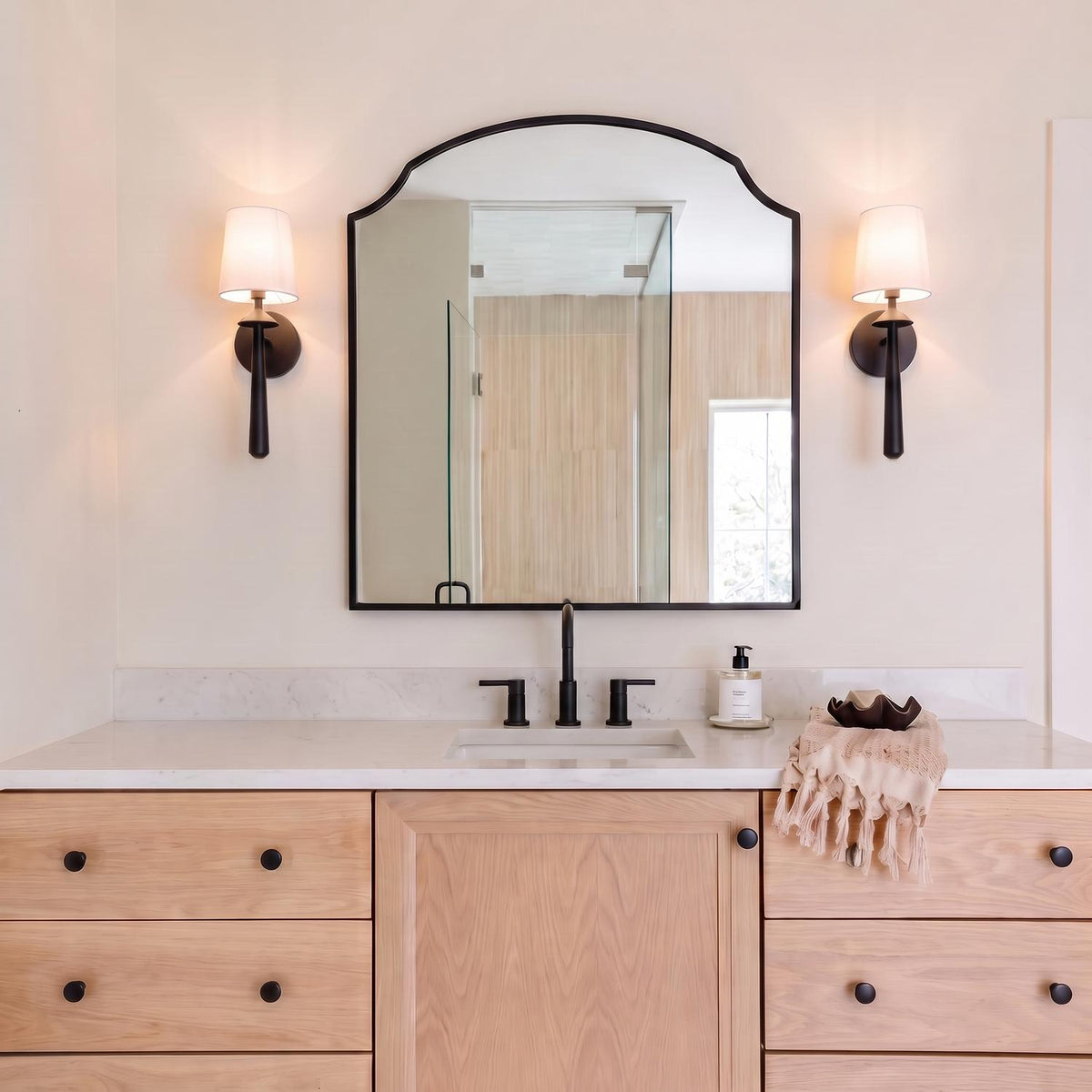 A transitional bathroom with two Sabina Wall Sconce 18", Matte Black wall sconces with white linen shades flanking an arched mirror above white marble and wood cabinetry.