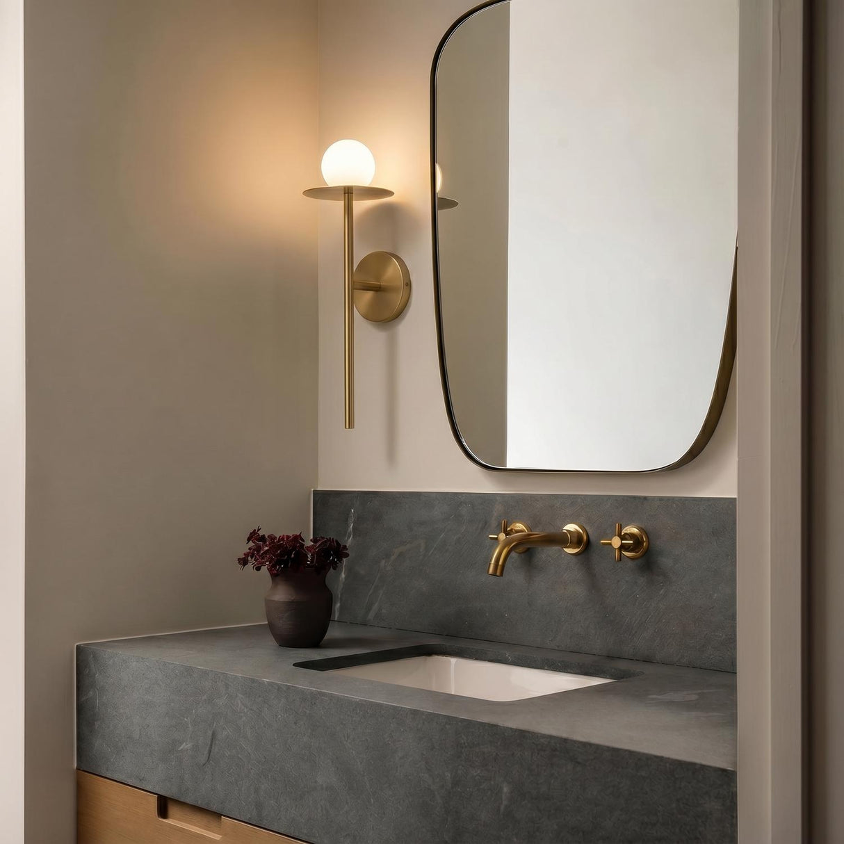 A modern bathroom with the Sol LED Wall Sconce 16", Brushed Brass featuring a frosted globe mounted beside a curved mirror over a dark gray soapstone vanity.