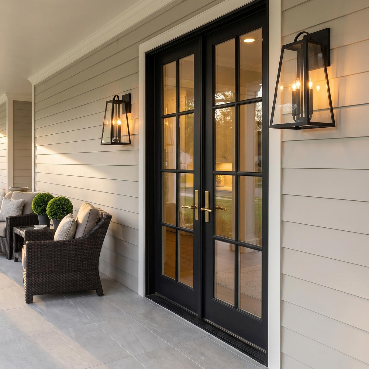 A modern farmhouse porch featuring two matte black Alden wall lanterns flanking black French doors on beige siding at dusk with wicker seating and boxwoods.
