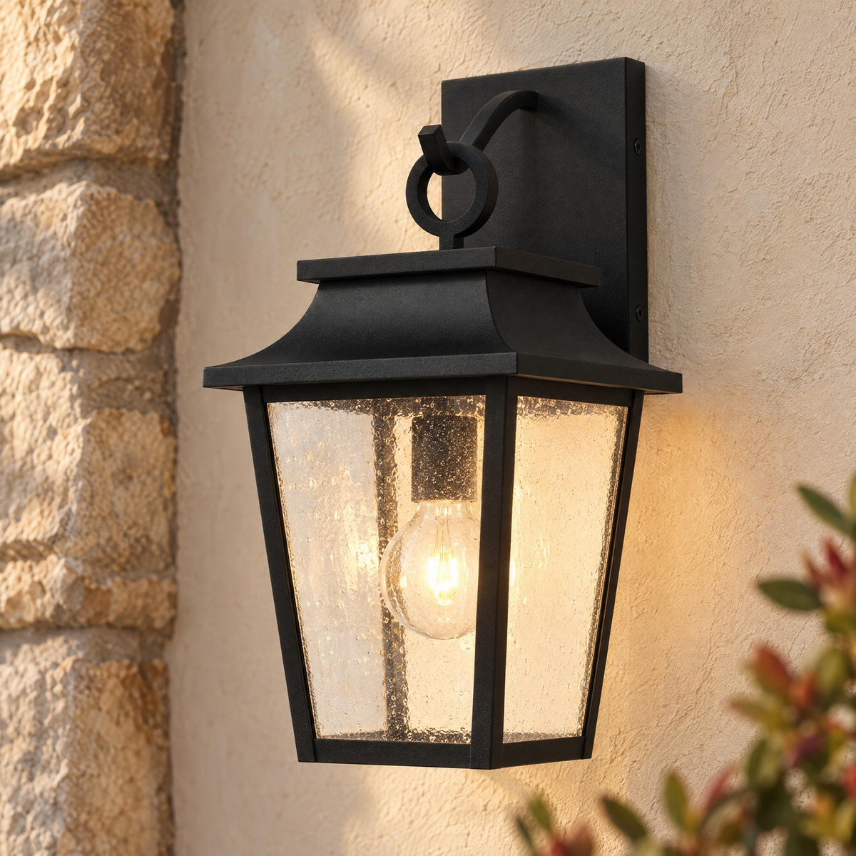A Mediterranean home exterior featuring a matte black coach lantern glowing on beige stucco beside a stacked stone column at dusk with greenery.