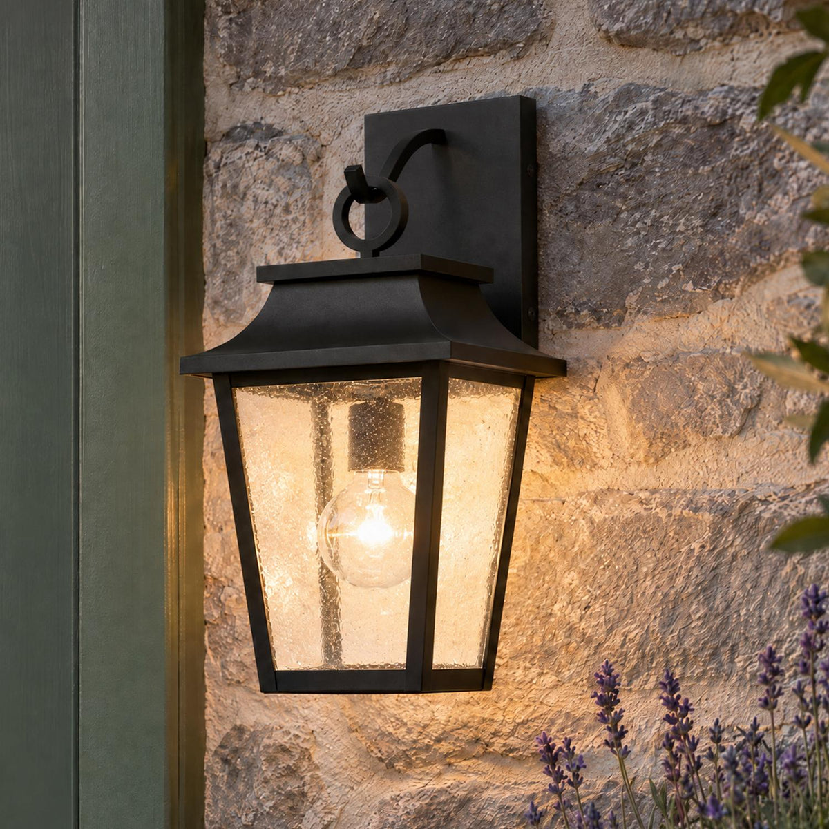A craftsman exterior features the glowing matte black Beckett Outdoor Wall Light, Small 16" Black mounted on a stone wall beside a green door with lavender.
