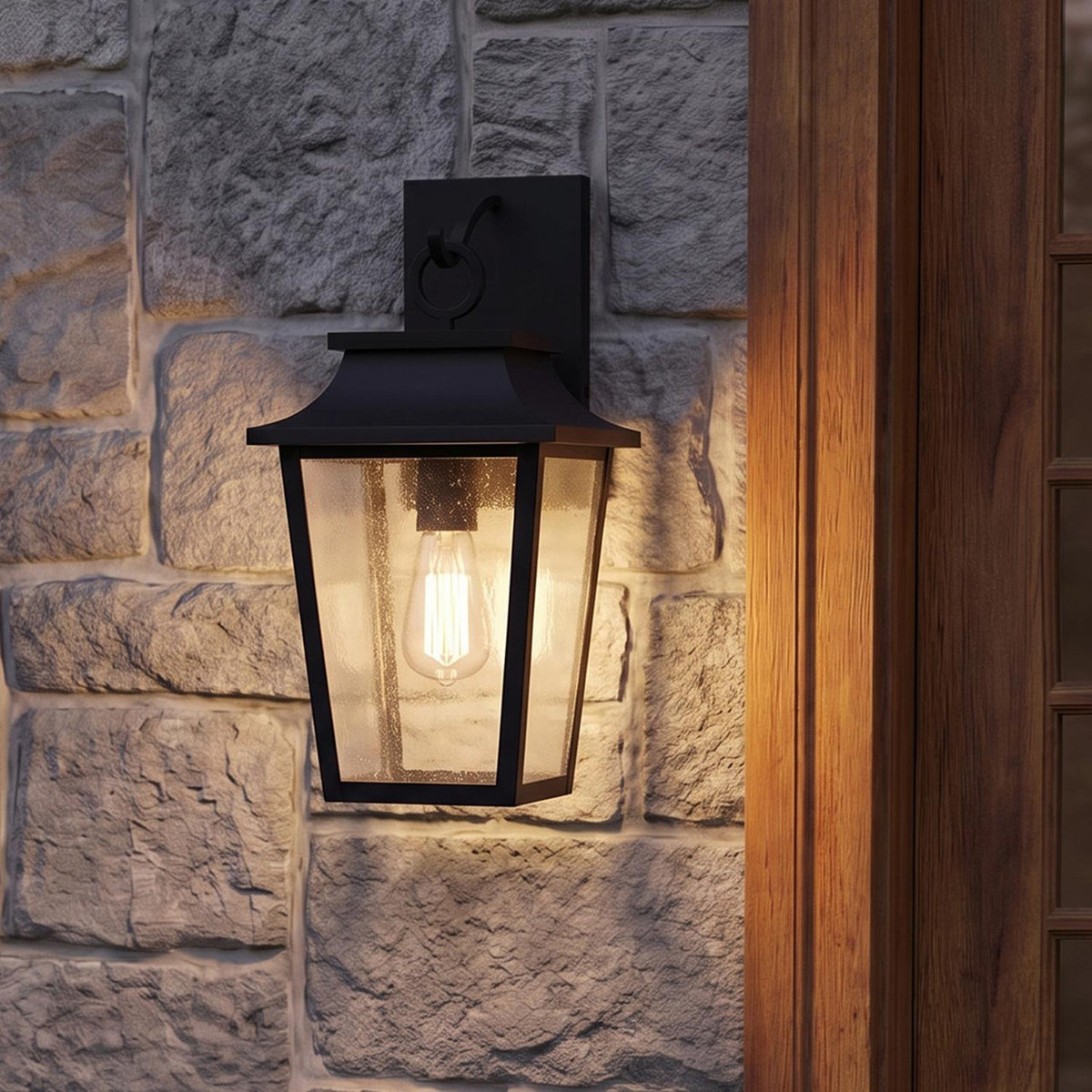 A craftsman home entry featuring the Beckett Outdoor Wall Light, Small 16" Black mounted on a gray stone wall beside a rich wood door at dusk.