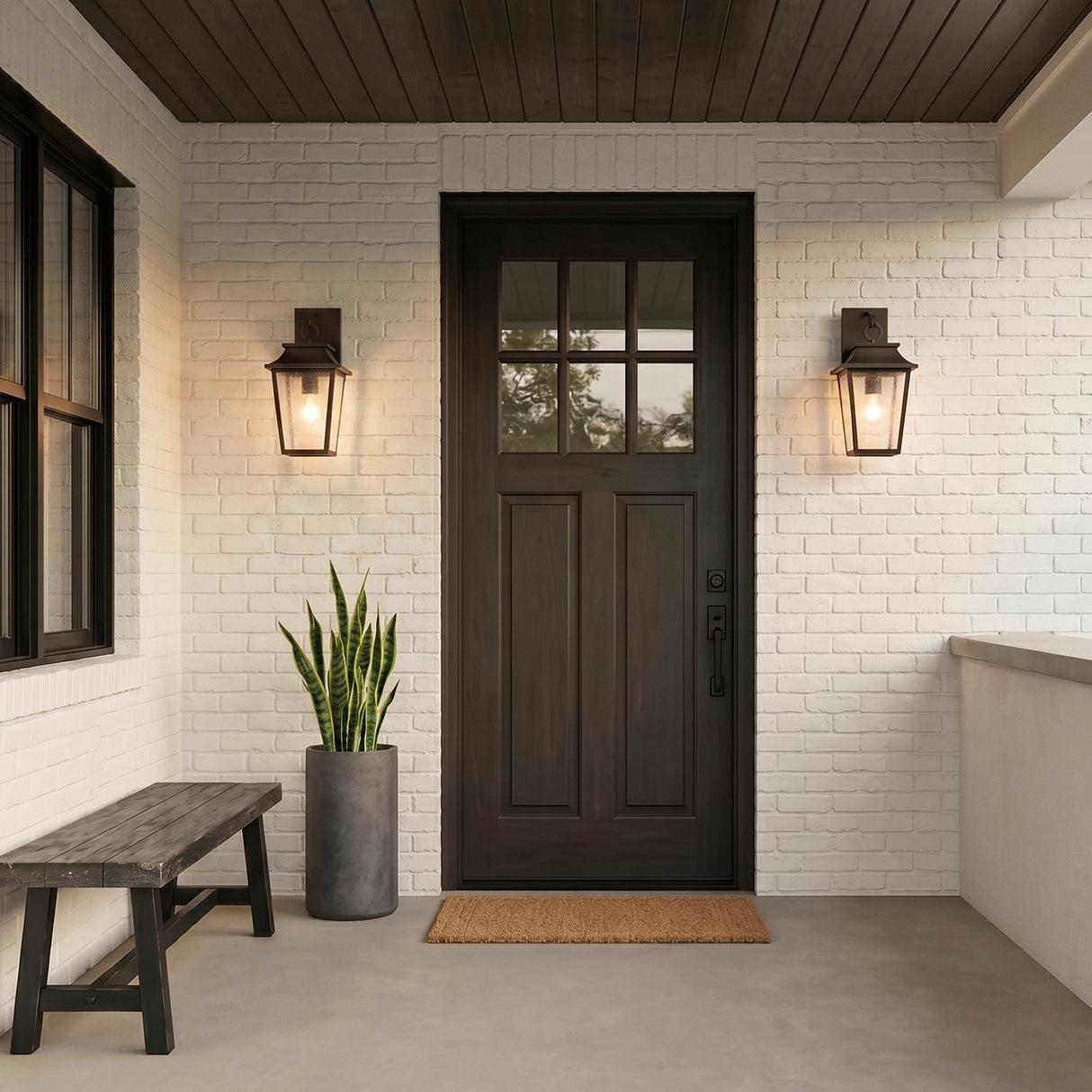 A modern farmhouse porch with two oiled bronze wall lanterns glowing at dusk, flanking a dark door on white brick beside a potted snake plant and bench.