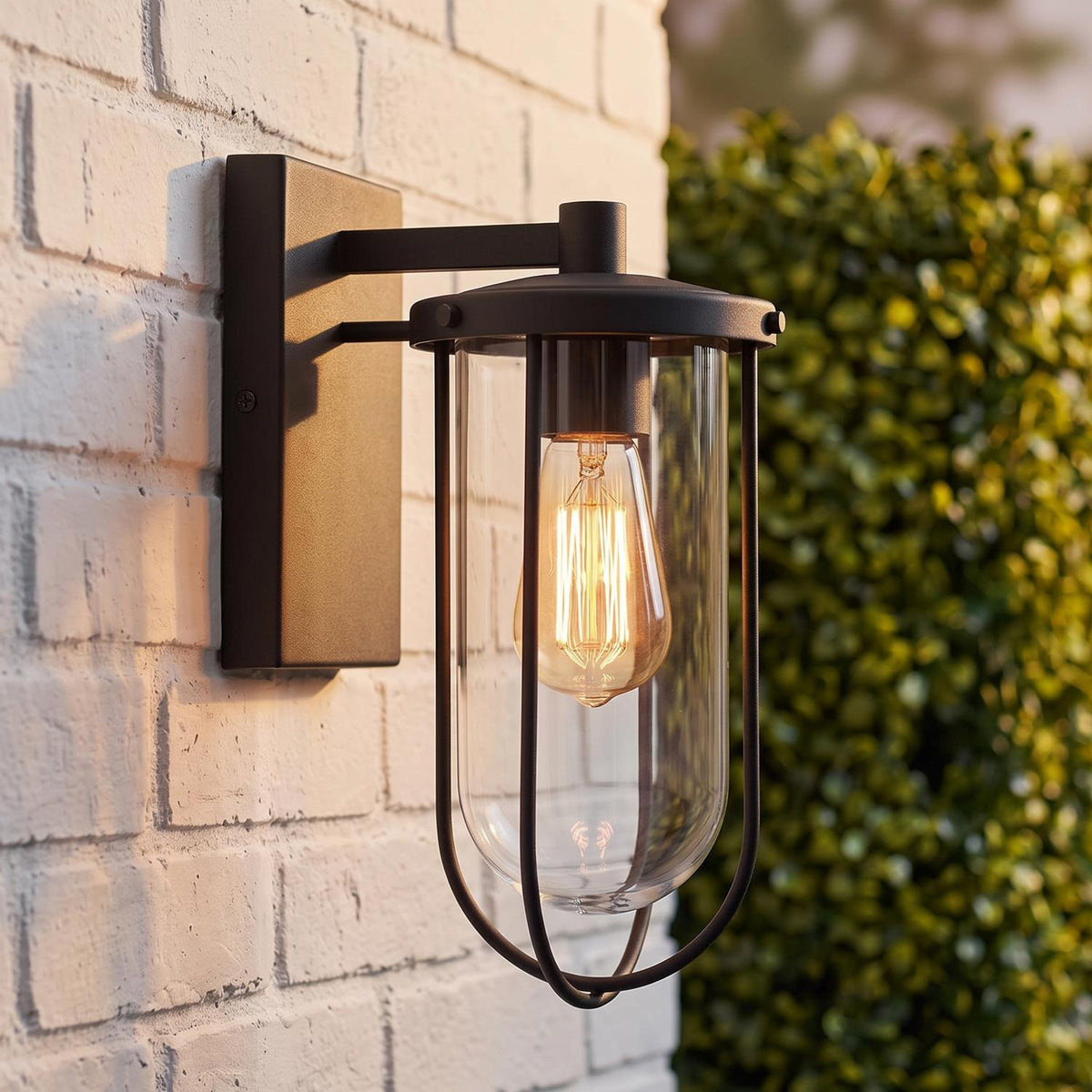 A modern farmhouse patio features the Orson Outdoor Wall Light, Small 12" Black, glowing in matte black against a white brick wall beside green hedges.