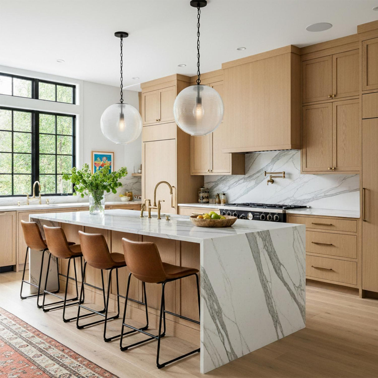 A modern kitchen features two Ori Pendant 17", Matte Black with ribbed glass over a marble waterfall island, complemented by light wood cabinets and leather barstools.