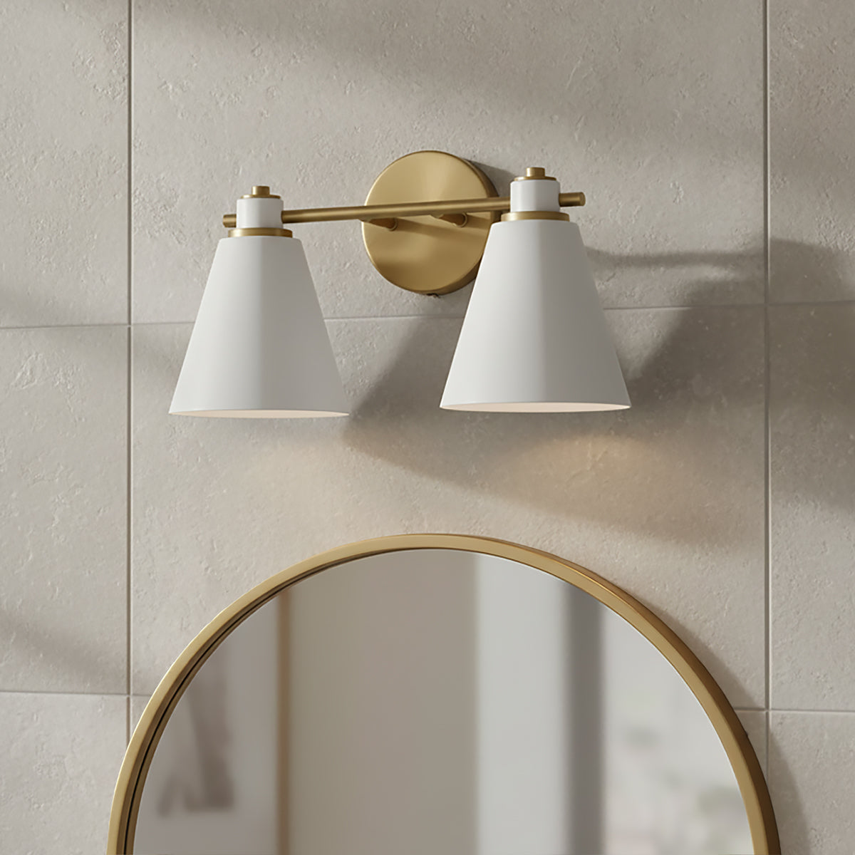 Avin 2 Light Vanity - Two white conical shades with brass accents on a brass bar, mounted above a round brass-framed mirror, against a tiled wall.