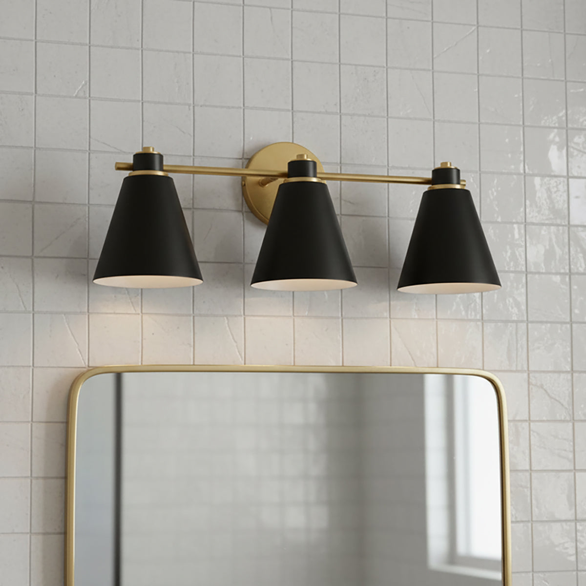 Avin 3 Light Vanity, Matte Black and Aged Brass - Three black conical shades with aged brass accents are mounted on a linear bar above a gold-framed mirror, all against a backdrop of white tiled walls.