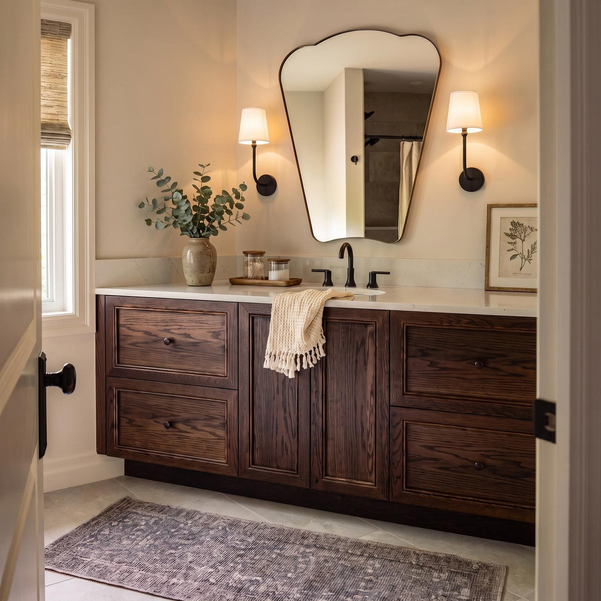 A transitional bathroom with two matte black Elowen Wall Sconces featuring linen shades flanking an organic-shaped mirror over a dark wood vanity and quartz countertop.