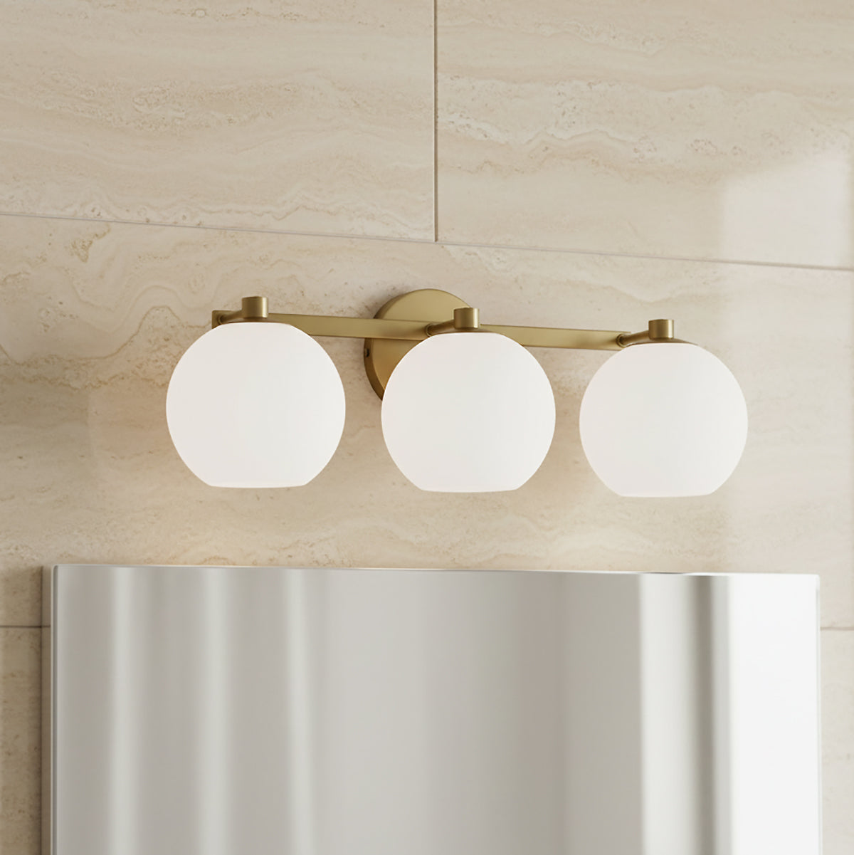 Enid 3 Light Vanity, Aged Brass - A three-globe vanity light fixture in aged brass with white glass shades is mounted on a travertine-tiled wall above a reflective mirror.