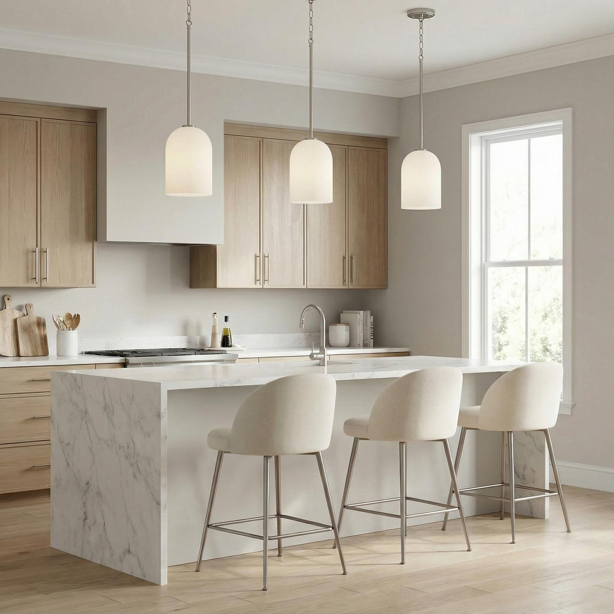 A contemporary kitchen featuring three Elira Pendant 9", Brushed Nickel fixtures with frosted glass shades over a white marble waterfall island and light wood cabinets.