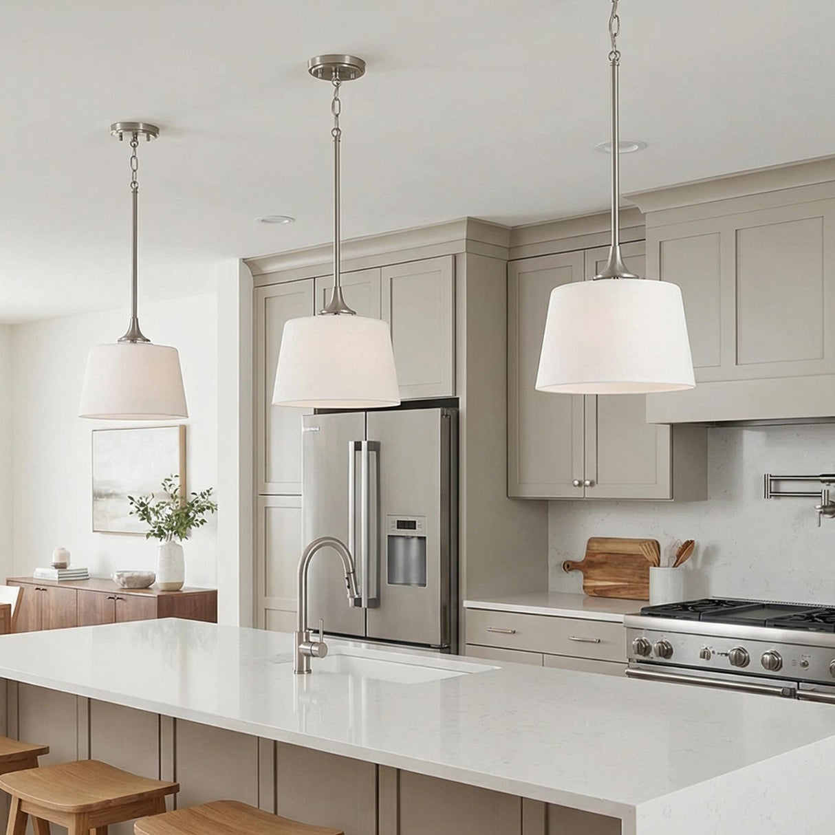 A transitional kitchen features three pendants with brushed nickel hardware and white glass drum shades installed over a white quartz island with taupe shaker cabinets.
