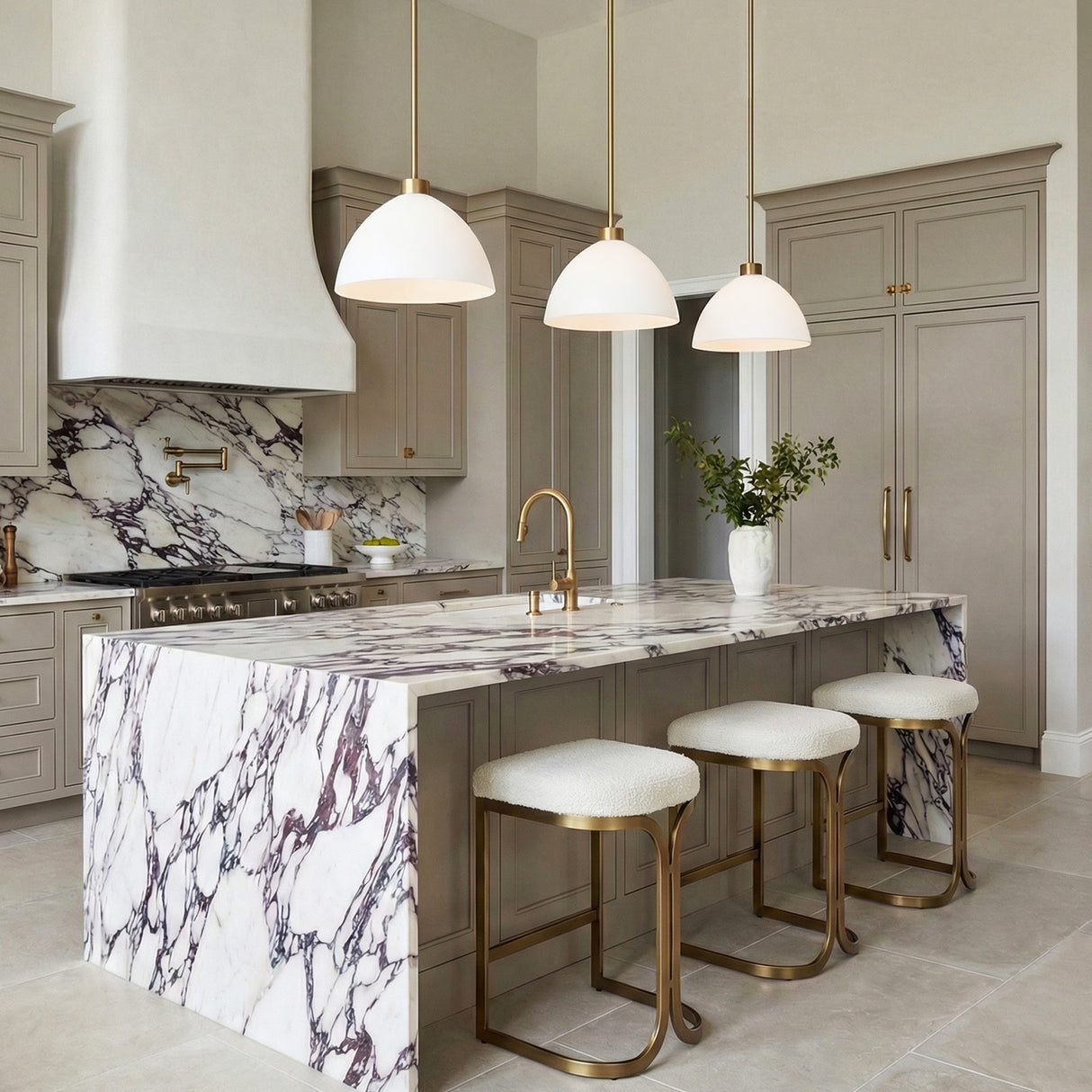 A transitional kitchen featuring three Cleo Pendant 13", White and Aged Brass fixtures over a purple-veined marble waterfall island with beige cabinets and a matching marble backsplash.