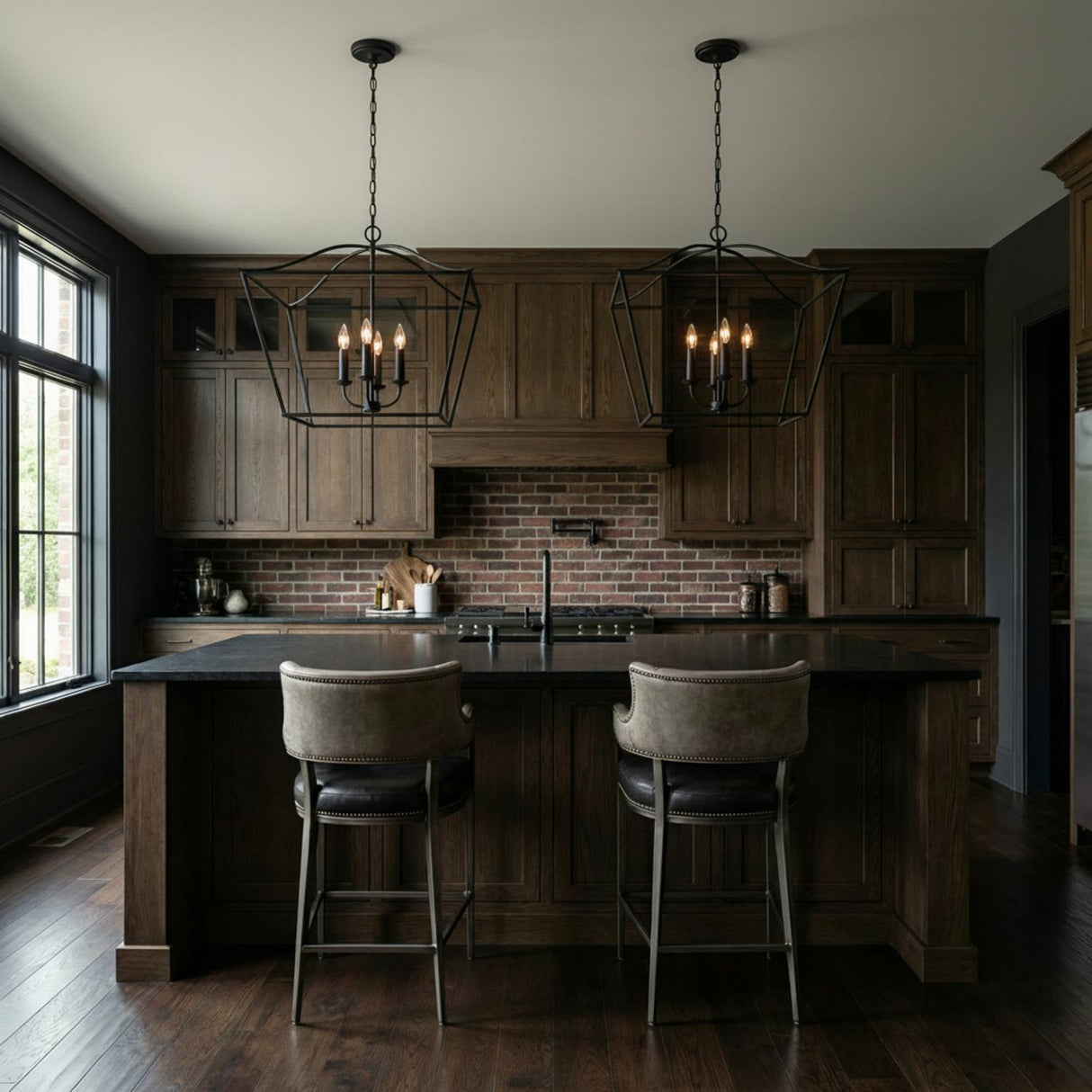A dark industrial kitchen features two Eloise Lantern Pendant 28", Textured Black fixtures over a black stone island paired with dark wood cabinets and a brick backsplash.