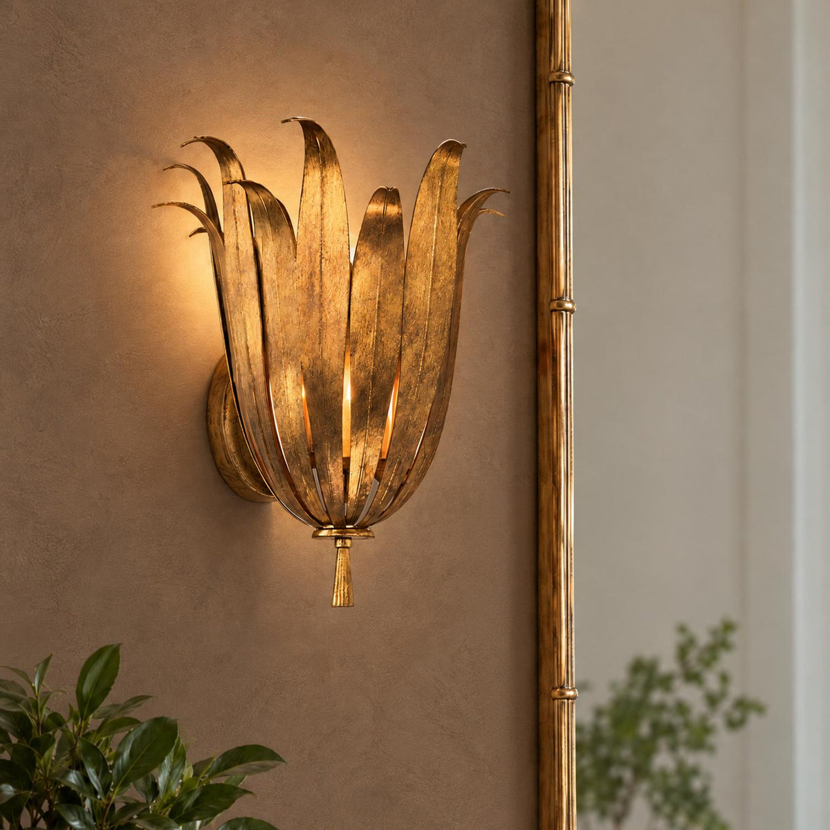 A transitional foyer features the Cerise Wall Sconce 14", Antique Gold mounted on a warm textured wall next to a gilded bamboo mirror and green plant.