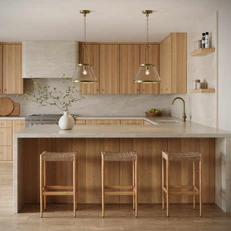 Two Elora 15" wide Pendants, in Aged Brass with Hammered Glass, hung above a kitchen peninsula with natural stone countertops and wood cabinets.
