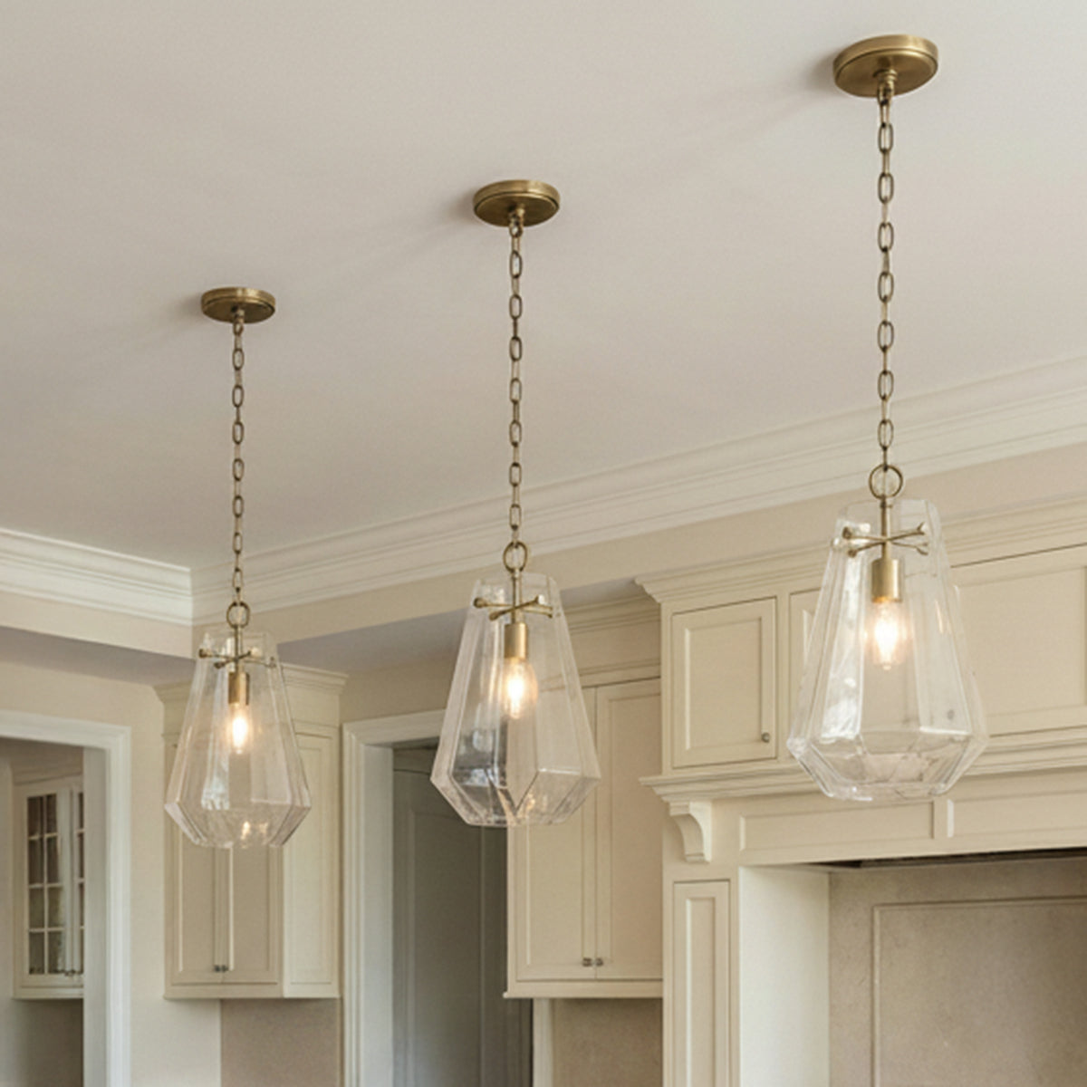 A traditional kitchen features three Delmar Pendant 11", Aged Brass fixtures with aged brass chains and faceted glass shades over the island alongside cream cabinetry.
