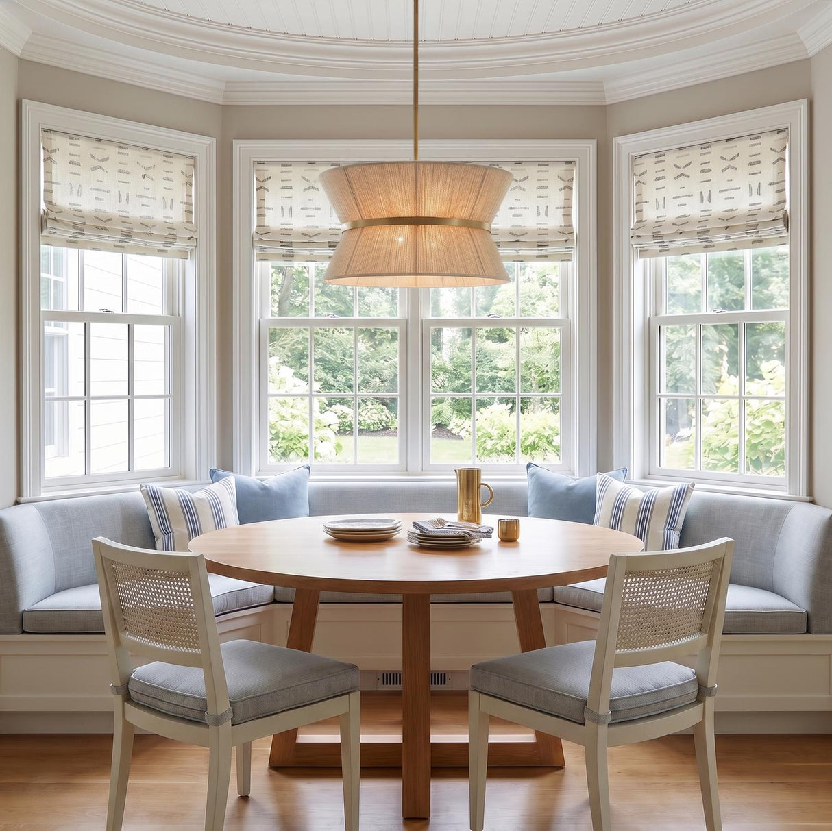 A coastal dining nook with a patina brass and linen drum pendant suspended over a round oak table with light blue banquette seating and white cane chairs.