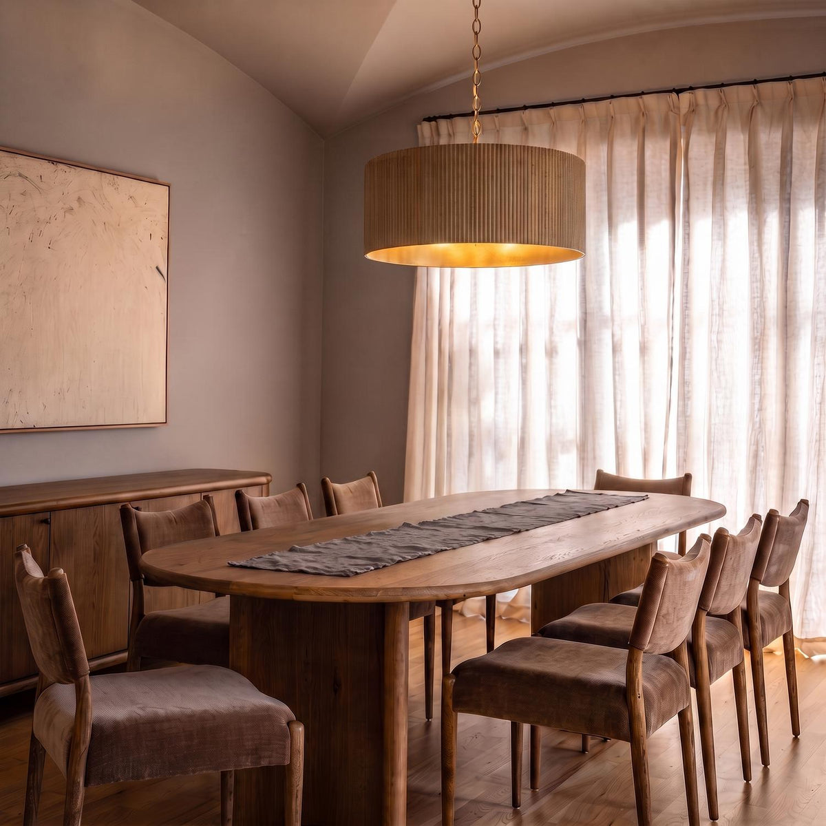 A contemporary dining room with a ribbed whitewash wood drum pendant featuring matte brass hardware hanging over an oval wood table under a vaulted ceiling.